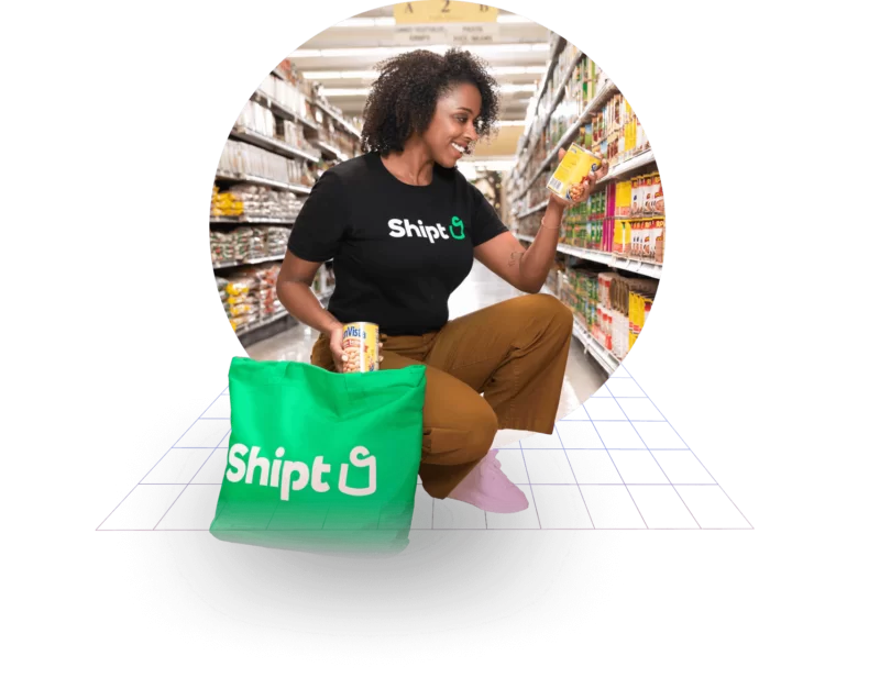 A woman crouched in a grocery store looking at a can of food while putting another can of food into a green Shipt bag.
