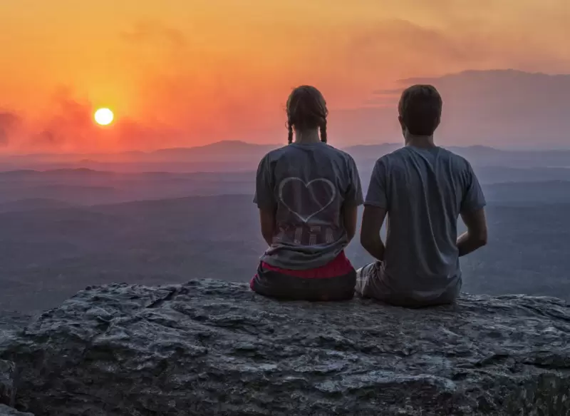 A couple sitting on the edge of a cliff watching the sun set.