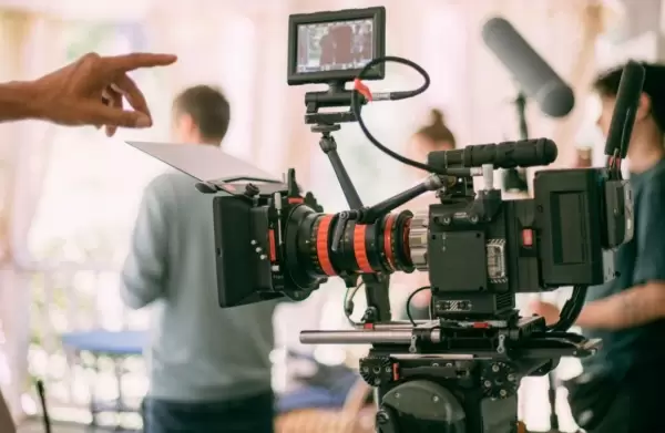 A video camera with various people working on set behind it.