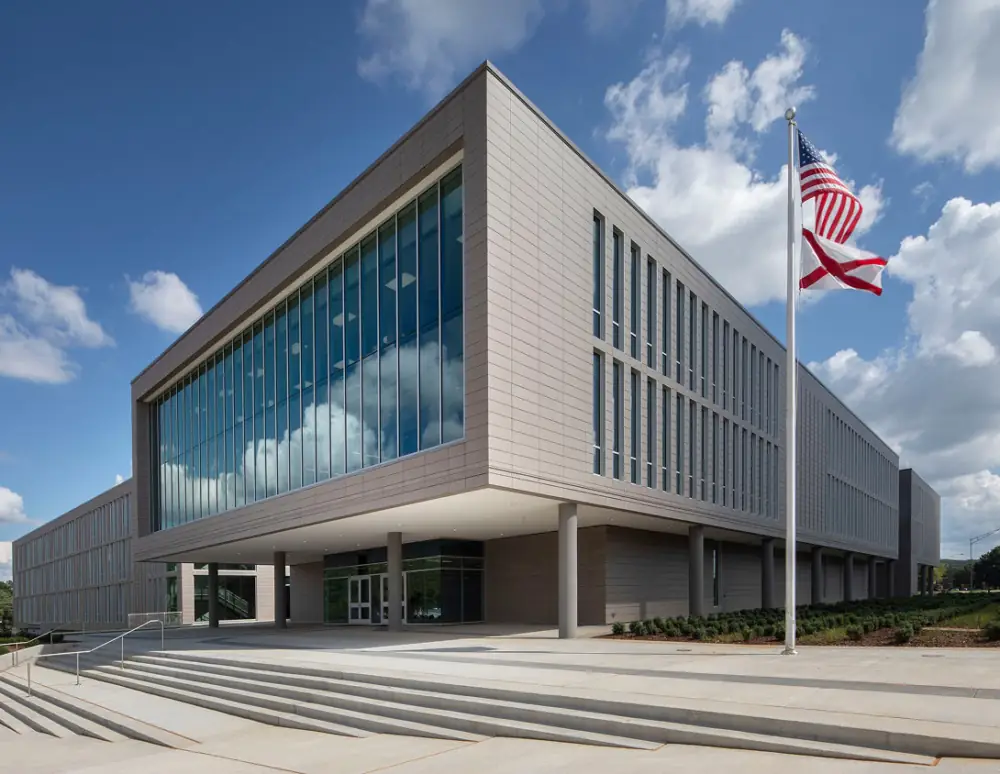 Exterior of a modern square building with full-height windows. The United States flag flies on a flagpole with the Alabama flag below it.