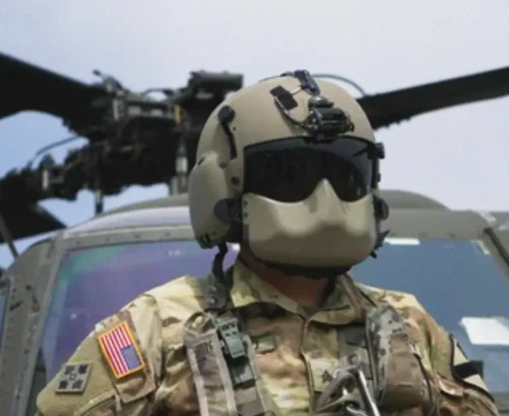 A helicopter pilot wearing a camouflage suit and flight helmet standing in front of a helicopter.